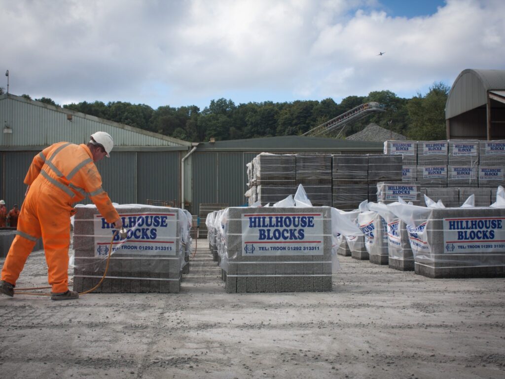 Person in safety gear examines Hillhouse Blocks at industrial site with sheds and conveyor.