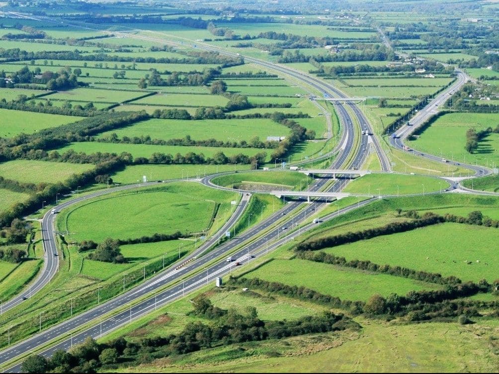 Aerial view of motorway interchange with roads, overpasses, green fields, trees, and rural landscape.
