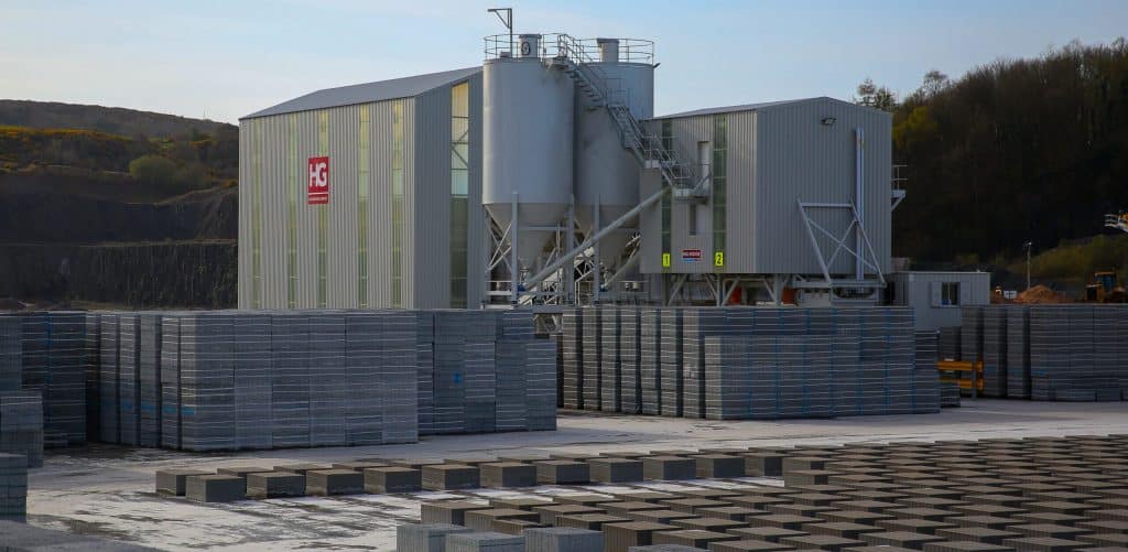 Industrial site with metal buildings, silos, concrete blocks, wooded landscape in background.