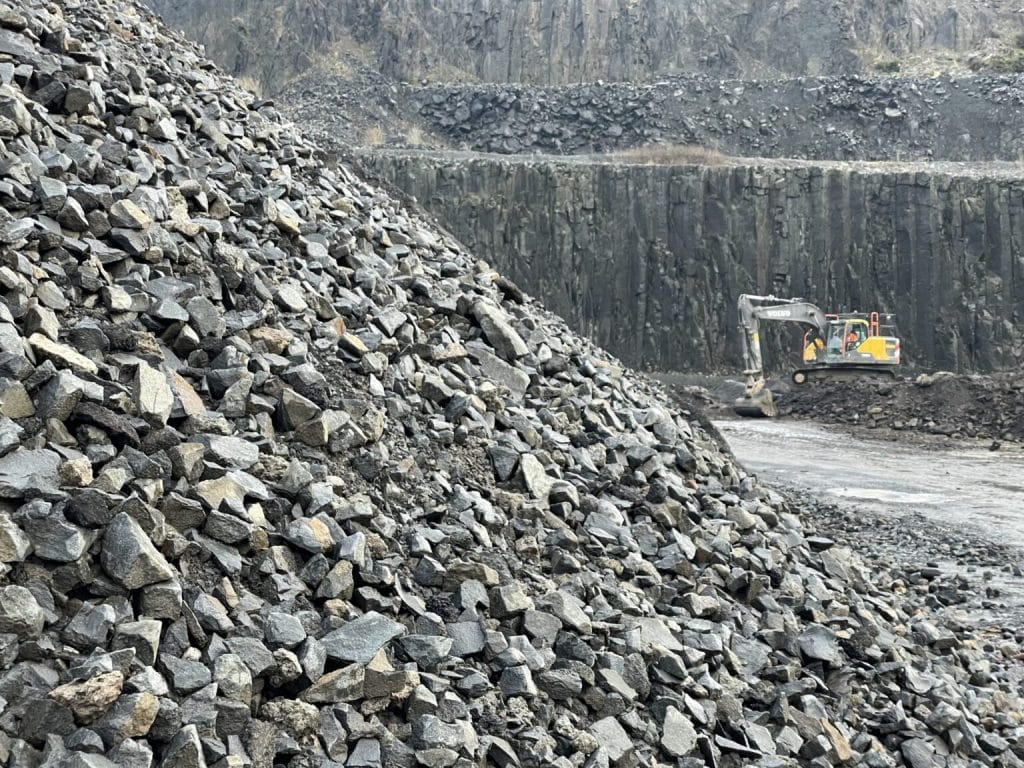 Quarry with rock piles and construction vehicle on dirt road against steep rock walls.