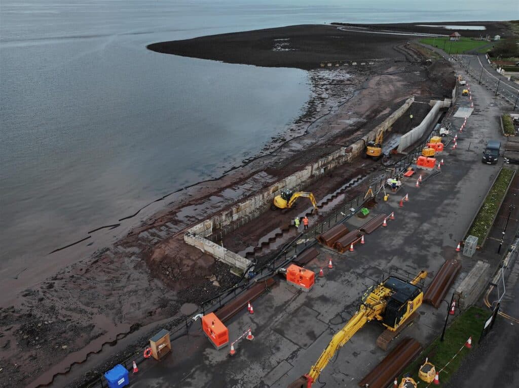 Coastal site with excavators, machinery, sea wall under construction, marked by cones and barriers.