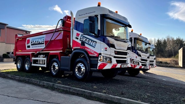 Two large trucks with "Skene" branding parked in a lot under a partly cloudy sky.