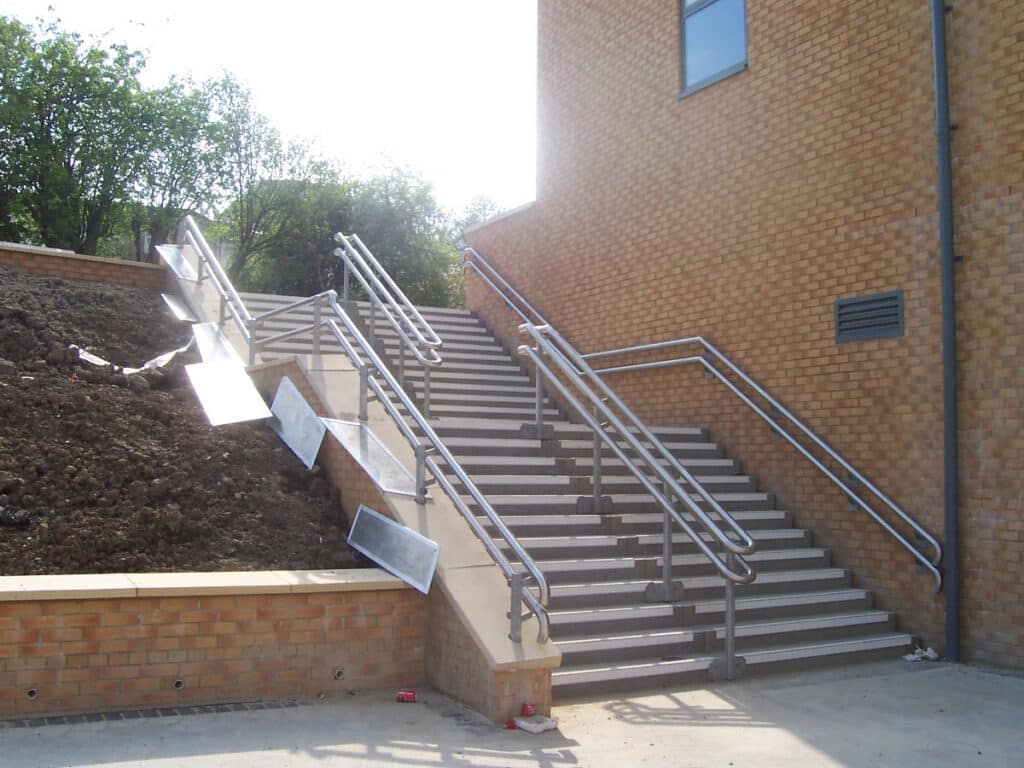 Outdoor concrete stairs with metal railings beside a brick building and a soil embankment.