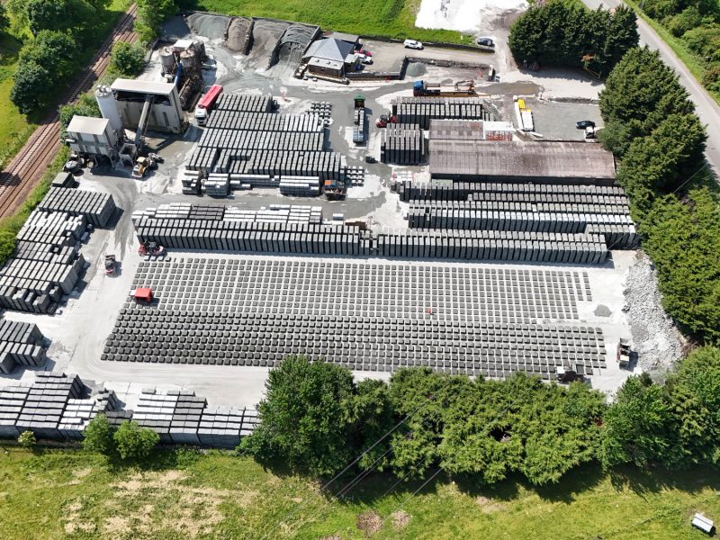 Aerial view of industrial site with stacked concrete blocks, machinery, buildings, road, and railway.