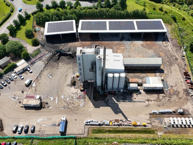 Aerial view of industrial site with silos, buildings, parking, solar panels, and surrounding greenery.
