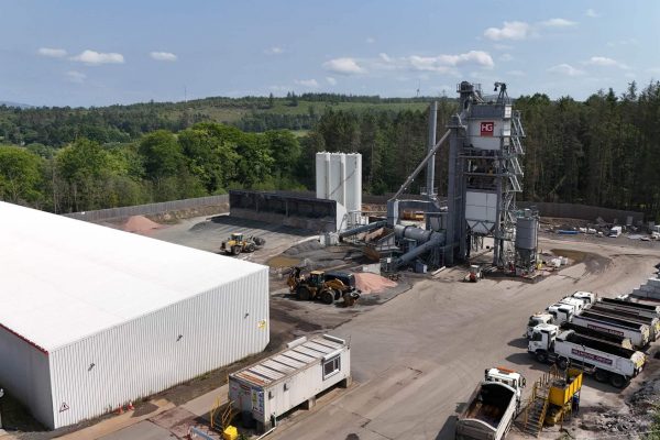 Aerial view of industrial site with warehouse, silos, construction vehicles, trucks, and surrounding trees.