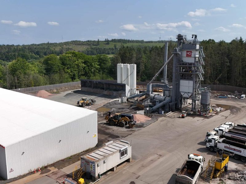 Aerial view of industrial site with warehouse, silos, construction vehicles, trucks, and surrounding trees.