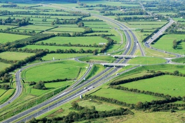 Aerial view of motorway interchange with roads, overpasses, green fields, trees, and rural landscape.