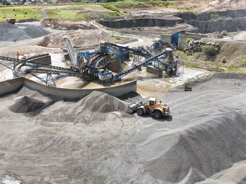 Aerial view of quarry with conveyor system, gravel piles, and yellow front loader.