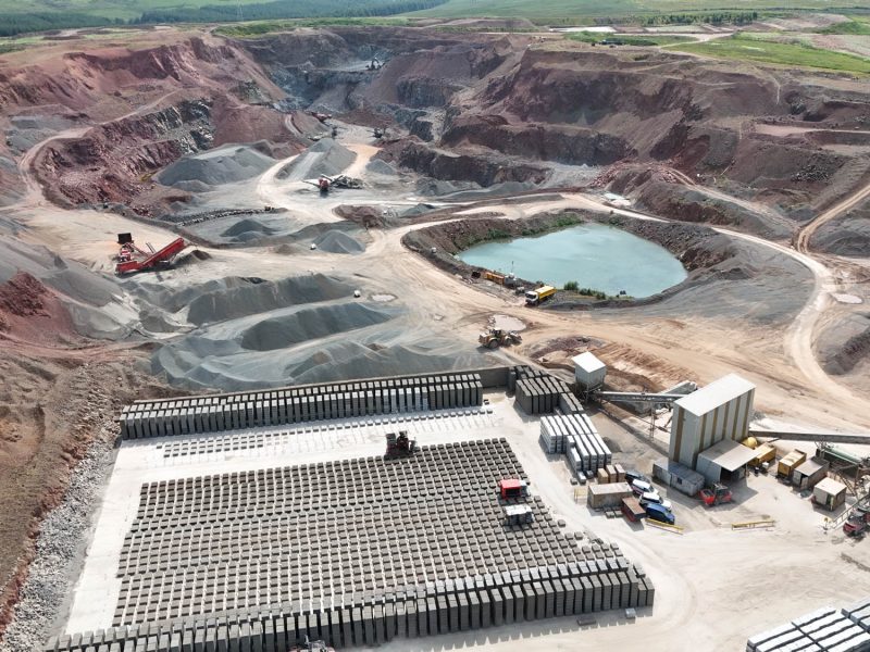 Aerial view of quarry with gravel piles, concrete blocks, water body, buildings and vehicles.