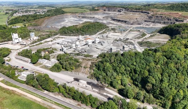 Aerial view of a quarry and industrial site with roads, buildings, forests, and nearby houses.