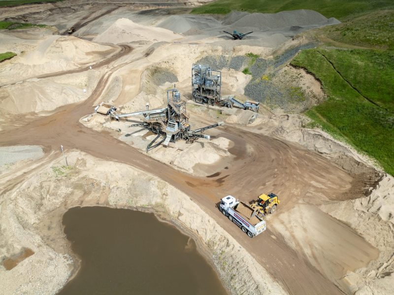 Aerial view of quarry with machinery, vehicles, sand piles, pond, and green landscape patches.