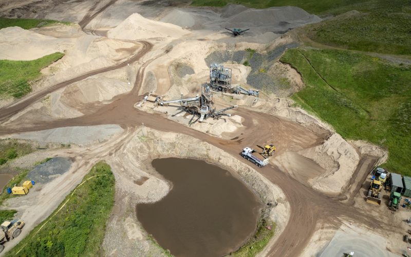 Aerial view of sand quarry with machinery, dirt roads, sand mounds, fields, and water body.