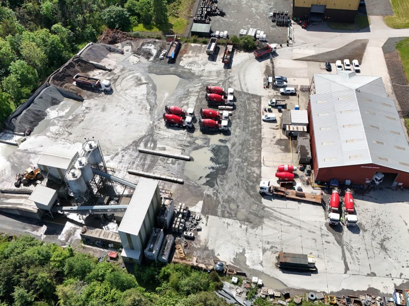 Aerial view of a concrete mixing plant with red mixer trucks and surrounding green trees.