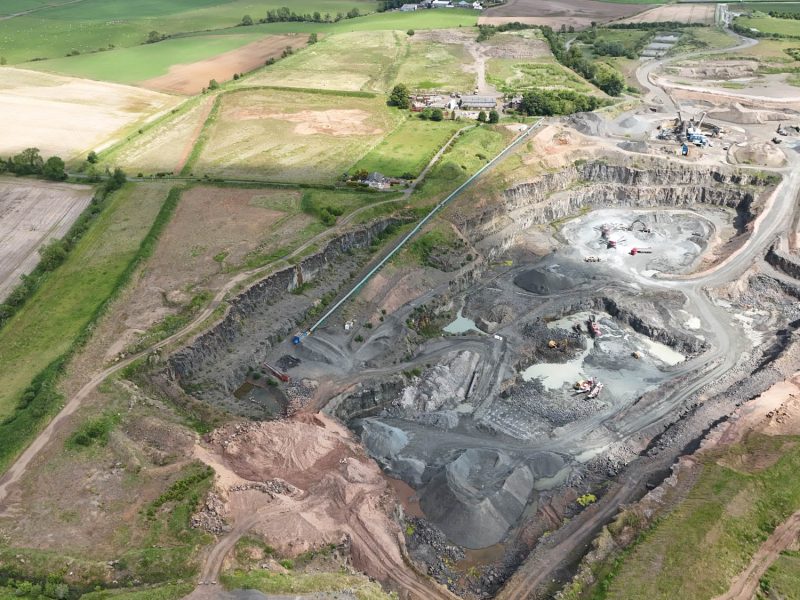 Aerial view of a multi-level quarry with machinery, piles of materials, fields, and nearby buildings.