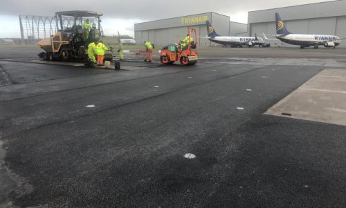 Airport tarmac resurfacing with workers, machinery, Ryanair planes, and hangars under overcast sky.