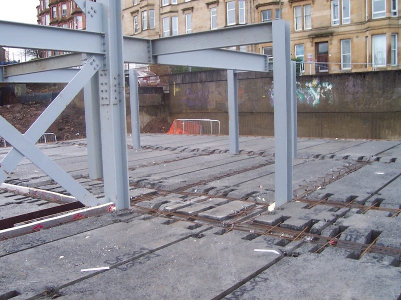 Construction site with steel framework, concrete slabs, graffiti-covered wall, urban buildings background.