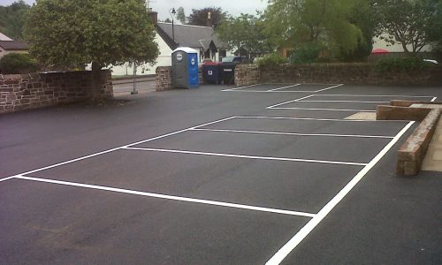 Empty outdoor car park with marked spaces, stone walls, trees, and a portable toilet visible.