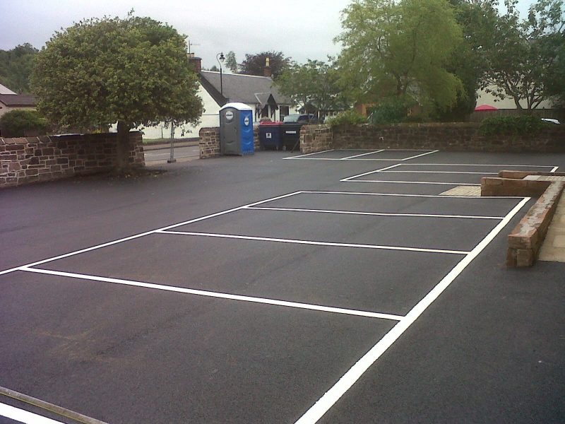 Empty outdoor car park with marked spaces, stone walls, trees, and a portable toilet visible.
