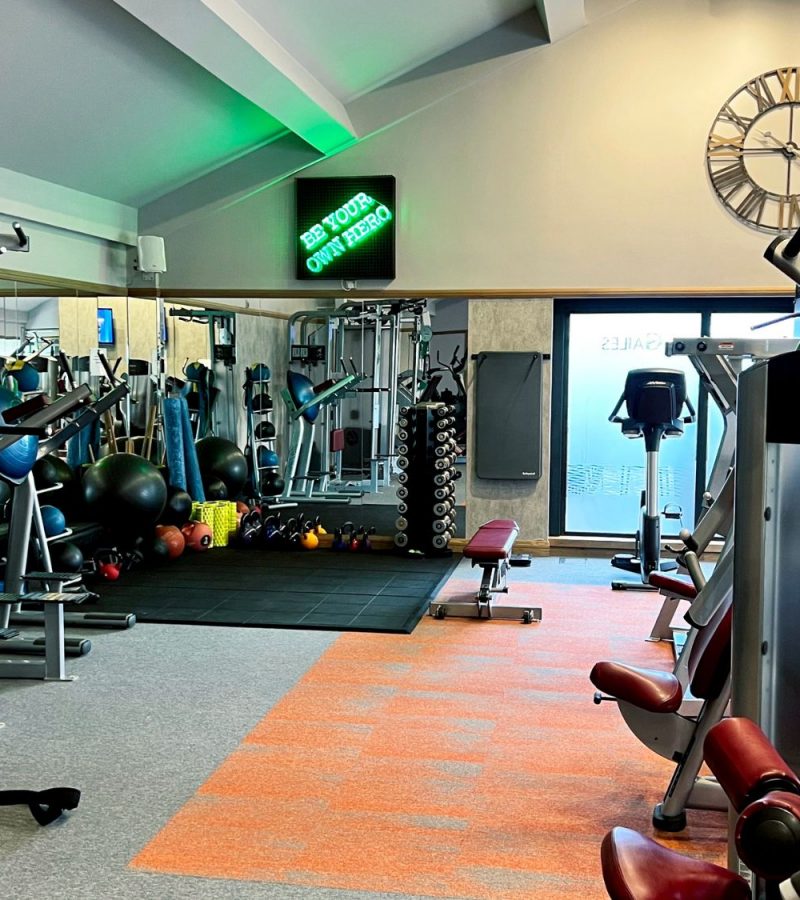 Gym with machines, weights, mirrored wall, motivational sign, clock, grey and orange flooring.