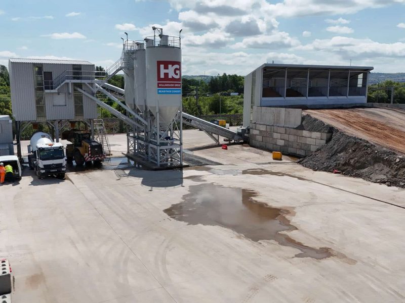 Industrial facility with concrete mixers, vehicles, concrete pavement, and earth pile nearby.
