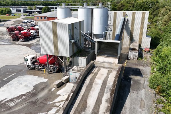 Industrial concrete plant with silos, conveyor belt, and red cement mixer trucks parked nearby.