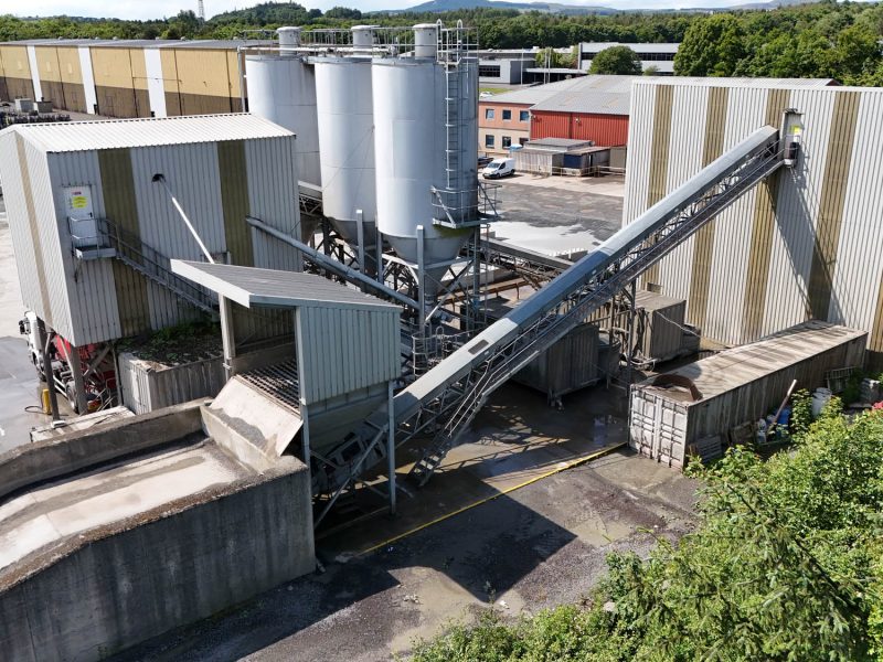 Industrial plant with storage silos, metal buildings, conveyor belts, and surrounding vegetation.