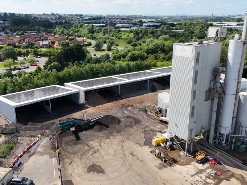 Industrial site with large white silos, covered materials, trees, and residential backdrop