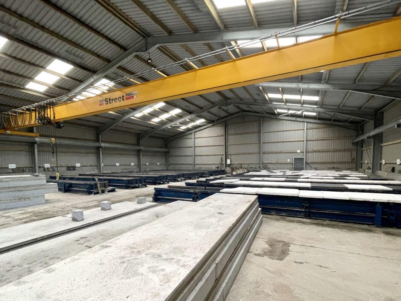 Industrial warehouse interior with yellow crane, concrete slabs on supports, skylit metal roof.