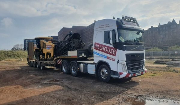 Large truck with low-loader trailer carrying machinery at a construction site under cloudy sky.