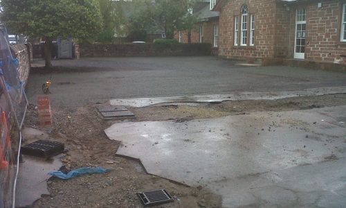 Partially excavated area with paving, drainage grates, tools, brick building, and tree in view.