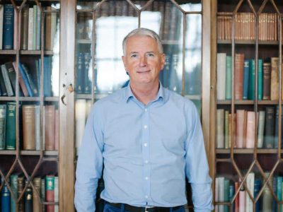 Person in blue shirt in front of a wooden bookshelf with glass doors filled with colourful books.