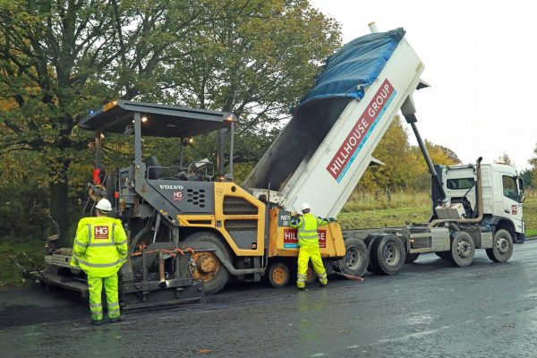 Workers operate road machinery; truck unloads materials into paver, near trees on tarmac.