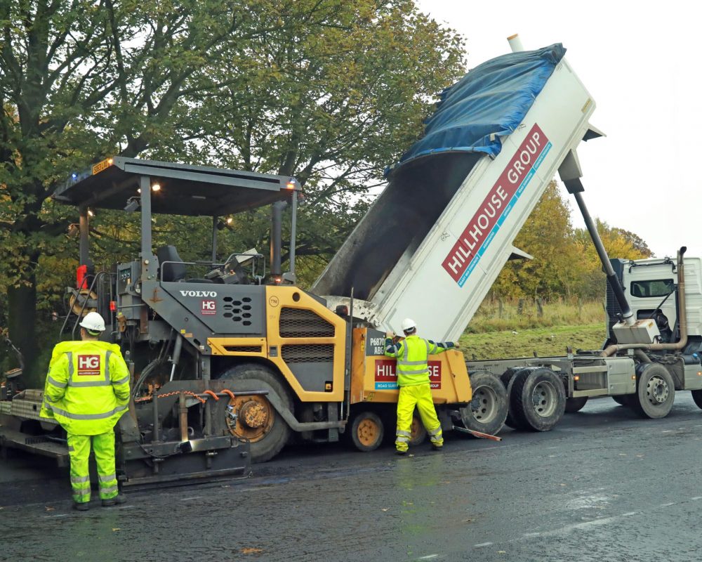 Workers operate road machinery; truck unloads materials into paver, near trees on tarmac.