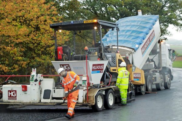 Road construction with workers in high-visibility gear paving asphalt, truck and trees behind.