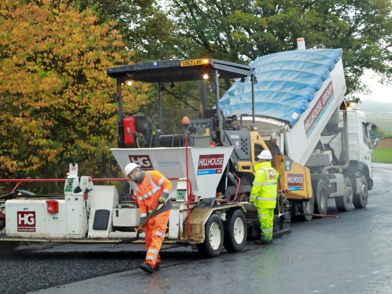 Road construction with workers in high-visibility gear paving asphalt, truck and trees behind.