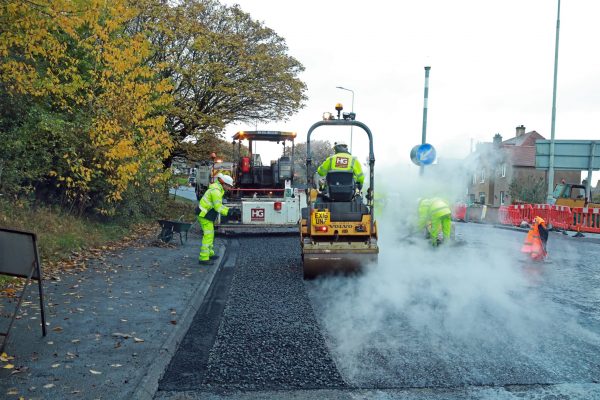 Road workers in high-vis clothing resurfacing road with machines, steam, and autumn trees.