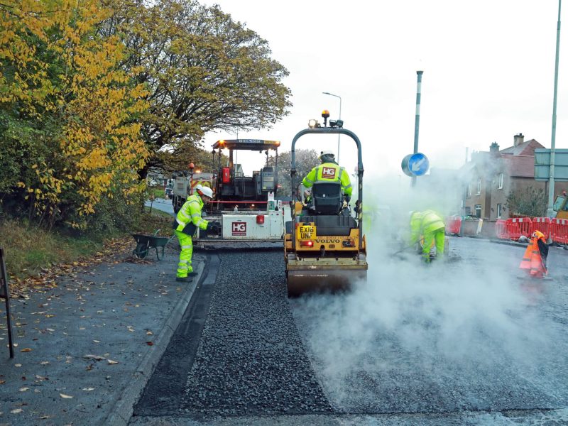 Road workers in high-vis clothing resurfacing road with machines, steam, and autumn trees.