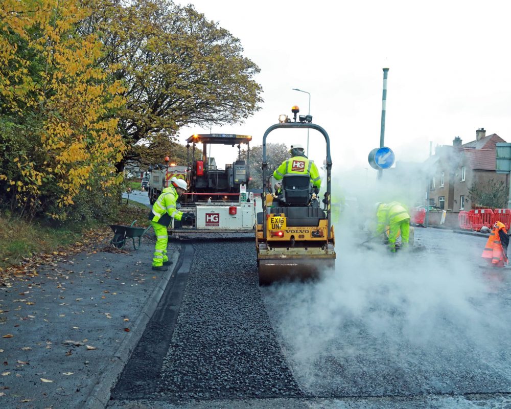Road workers in high-vis clothing resurfacing road with machines, steam, and autumn trees.
