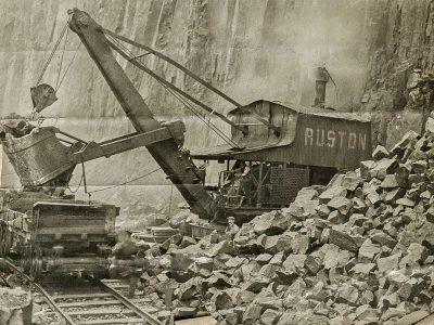 Mechanical excavator labeled "Ruston" loads rocks onto railcar in quarry with rocky cliff.