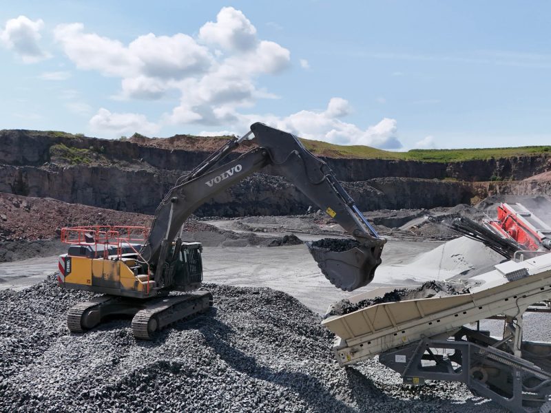 Volvo excavator loading crushed rocks onto conveyor belt in a rocky open quarry under blue sky.