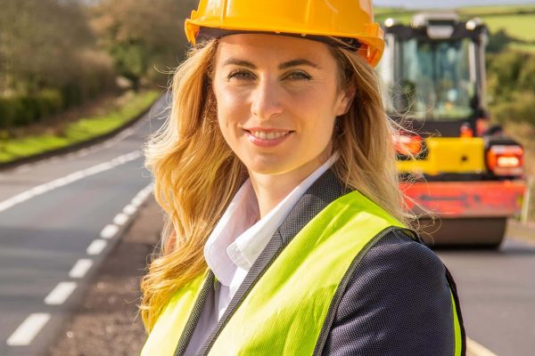 Woman in hard hat and high-vis vest at rural road construction site with a road roller behind.