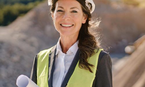 Woman in hard hat and high-vis vest holding a document at a construction site.