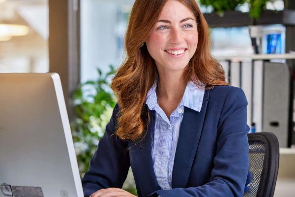 Woman with red hair in blue suit working at a computer in an office with plants and documents.