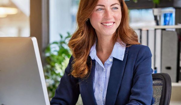 Woman with auburn hair in blue blazer, smiling at desk with computer, office folders, and plants.